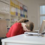A tired woman in a red sweater leans her head on a desk with a laptop, symbolizing workplace fatigue.