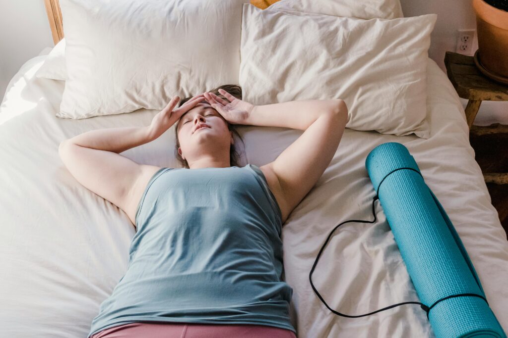 Woman lying on bed with a yoga mat beside her, appearing relaxed and comfortable indoors.