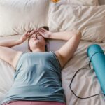 Woman lying on bed with a yoga mat beside her, appearing relaxed and comfortable indoors.