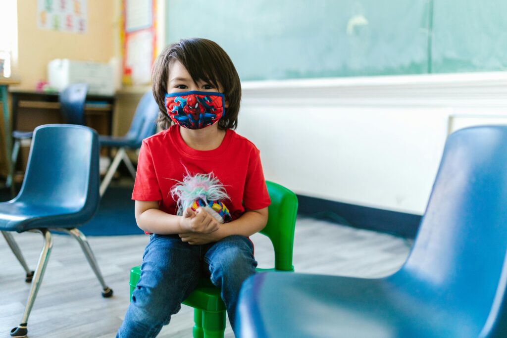 Child sitting in a classroom wearing a protective mask, surrounded by colorful chairs.