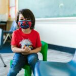 Child sitting in a classroom wearing a protective mask, surrounded by colorful chairs.